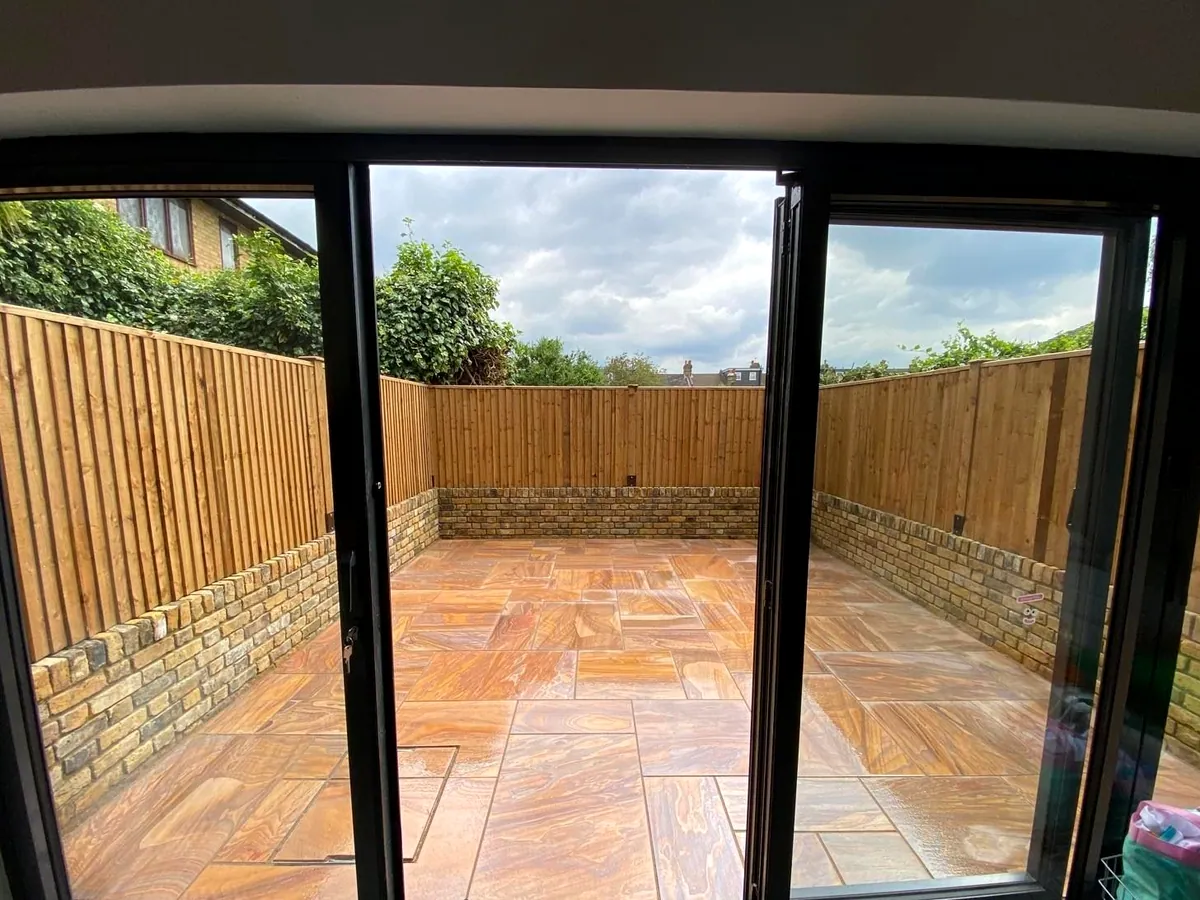Rainbow multicolour sandstone patio seen through open black bi-fold doors, with brick retaining walls and a feather-edge fence