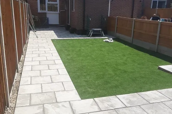 Cream and grey riven paving framing an artificial-grass lawn, with a feather-edge fence and red-brick house