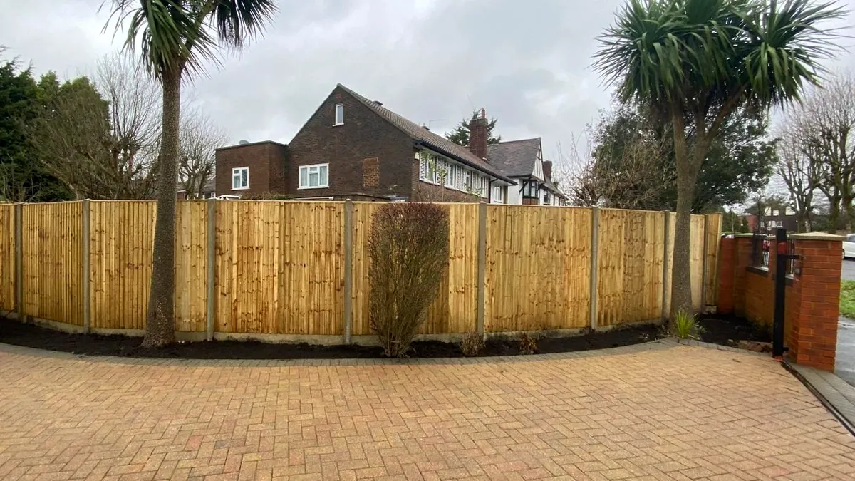 Feather-edge fence curved around a front garden between two cordyline palms, over a block-paved drive