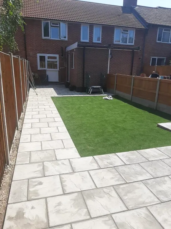 Cream and grey riven paving framing an artificial-grass lawn, with a feather-edge fence and red-brick house